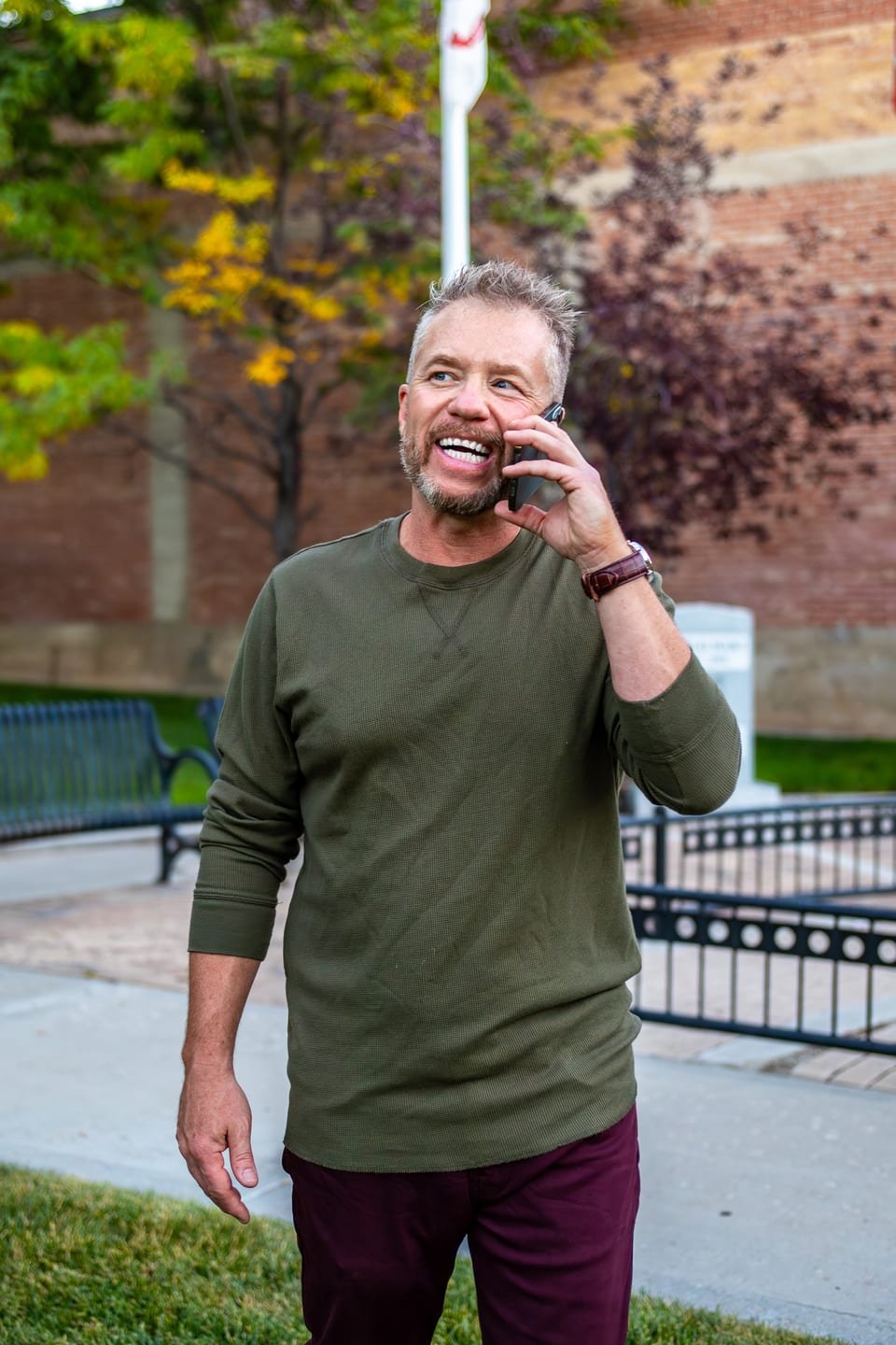 Cecil Hawthorne outdoors on Main Street, mid-call and laughing, in an olive henley — brick storefront and autumn trees behind him.